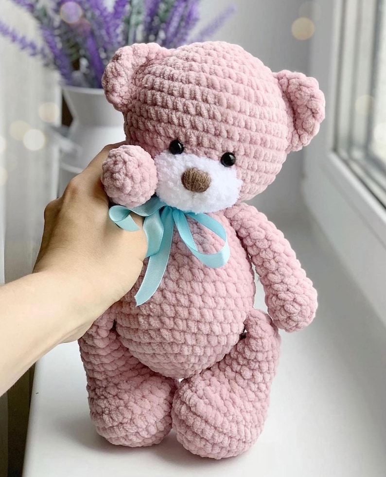 May include: A pink crocheted teddy bear with a white face and blue ribbon. The bear has black eyes and a brown nose.