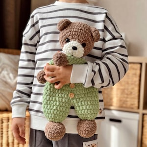 May include: A child holding a brown and green crocheted teddy bear wearing a green jumpsuit. The teddy bear has white eyes and a white nose.