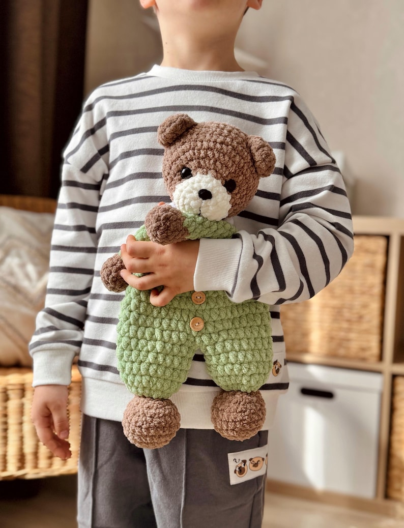 May include: A child holding a brown and green crocheted teddy bear wearing a green jumpsuit. The teddy bear has white eyes and a white nose.