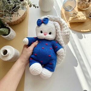 Puede incluir: Una mu&ntilde;eca de conejo hecha a mano con ganchillo. El conejo es blanco con orejas largas y ca&iacute;das y un lazo azul. Lleva un mono azul con detalles de corazones rojos. La mu&ntilde;eca es suave y afelpada, perfecta para un juguete infantil o decoraci&oacute;n de guarder&iacute;a.