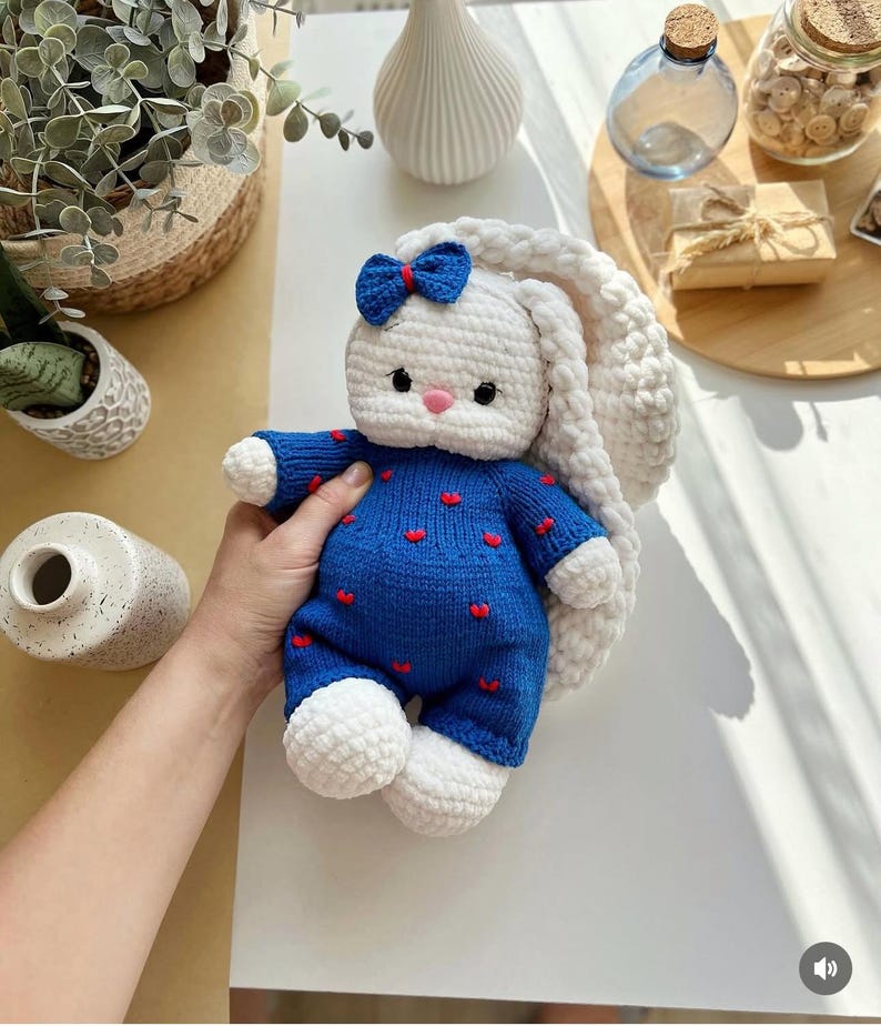 Puede incluir: Una mu&ntilde;eca de conejo hecha a mano con ganchillo. El conejo es blanco con orejas largas y ca&iacute;das y un lazo azul. Lleva un mono azul con detalles de corazones rojos. La mu&ntilde;eca es suave y afelpada, perfecta para un juguete infantil o decoraci&oacute;n de guarder&iacute;a.