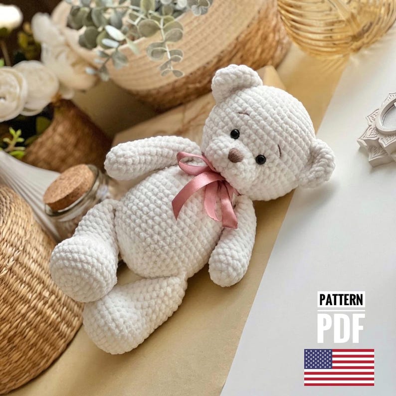 May include: White crocheted teddy bear with a pink ribbon around its neck. The bear has black eyes and a brown nose. The bear is lying on a light brown surface. The text "PATTERN PDF" is in the lower right corner of the image. An American flag is below the text.