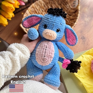 Pode incluir: Um brinquedo de pelúcia de burro azul feito à mão com orelhas rosa, barriga rosa e crina e cauda pretas. O brinquedo está nas mãos de uma pessoa. O texto "Pattern crochrt English" é visível.