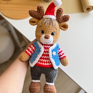 May include: A handmade crochet reindeer doll wearing a red and white striped shirt, a blue vest, and a Santa hat. The doll has brown antlers, gray pants, and brown shoes. The doll is a festive holiday decoration.