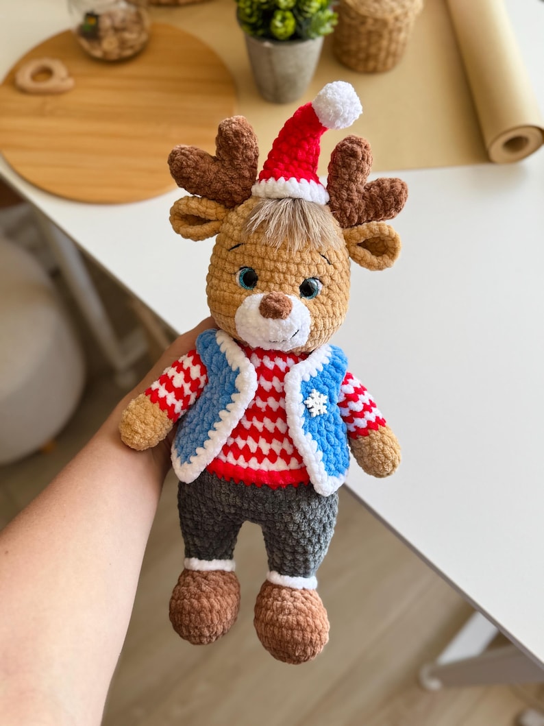 May include: A handmade crochet reindeer doll wearing a red and white striped shirt, a blue vest, and a Santa hat. The doll has brown antlers, gray pants, and brown shoes. The doll is a festive holiday decoration.