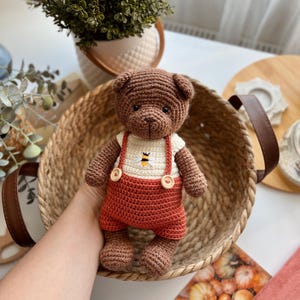May include: A handmade brown crochet teddy bear wearing a white shirt with a bee design and rust-colored overalls. The bear is in a woven basket with brown handles. The bear is a cute, soft toy.