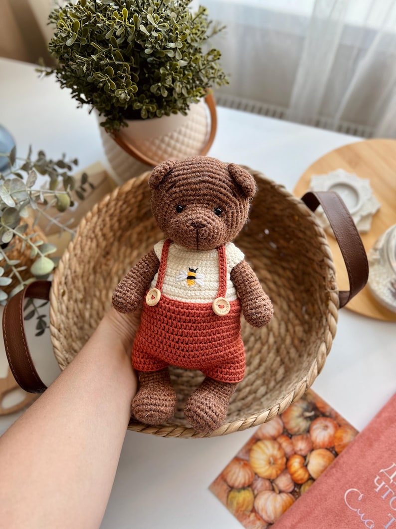 May include: A handmade brown crochet teddy bear wearing a white shirt with a bee design and rust-colored overalls. The bear is in a woven basket with brown handles. The bear is a cute, soft toy.