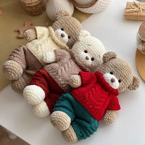 May include: A collection of handmade crochet teddy bears in various colors. Each bear wears a sweater and pants in shades of cream, brown, red, and teal. The bears have small black eyes and brown noses. The toys are arranged on a white surface.
