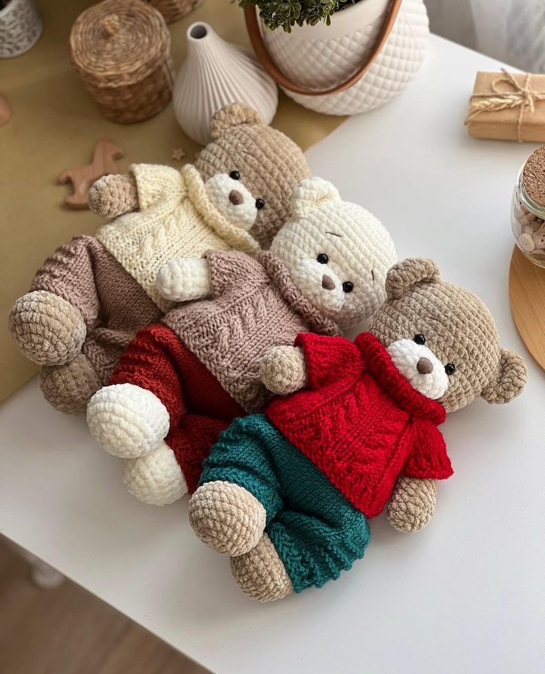 May include: A collection of handmade crochet teddy bears in various colors. Each bear wears a sweater and pants in shades of cream, brown, red, and teal. The bears have small black eyes and brown noses. The toys are arranged on a white surface.