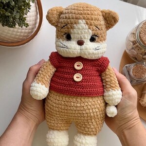 Pu&ograve; includere: Bambola a forma di gatto all'uncinetto fatta a mano con un maglione rosso. Il gatto ha una combinazione di colori marrone e bianco, con viso e zampe bianche. Il maglione rosso ha due bottoni di legno. La bambola &egrave; tenuta da una persona.