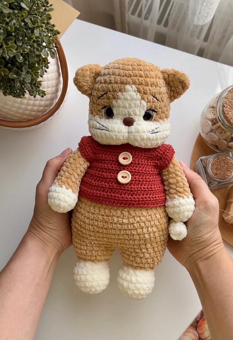 Pu&ograve; includere: Bambola a forma di gatto all'uncinetto fatta a mano con un maglione rosso. Il gatto ha una combinazione di colori marrone e bianco, con viso e zampe bianche. Il maglione rosso ha due bottoni di legno. La bambola &egrave; tenuta da una persona.