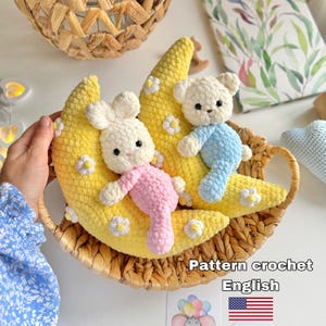 Pode incluir: Dois brinquedos de pelúcia em crochê, um coelho branco com detalhes rosa e um urso branco com detalhes azuis, descansando sobre uma almofada amarela em forma de meia-lua decorada com pequenas flores brancas em uma cesta de vime.