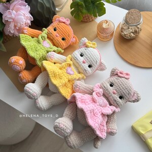 Pode incluir: Quatro bonecas de gato de croch&ecirc; feitas &agrave; m&atilde;o em laranja, branco, amarelo e rosa. Cada gato usa um vestido e um la&ccedil;o. As bonecas est&atilde;o em uma superf&iacute;cie branca. As bonecas s&atilde;o feitas &agrave; m&atilde;o e t&ecirc;m tra&ccedil;os faciais bordados.