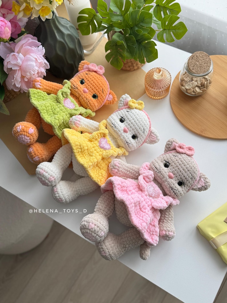 Pode incluir: Quatro bonecas de gato de croch&ecirc; feitas &agrave; m&atilde;o em laranja, branco, amarelo e rosa. Cada gato usa um vestido e um la&ccedil;o. As bonecas est&atilde;o em uma superf&iacute;cie branca. As bonecas s&atilde;o feitas &agrave; m&atilde;o e t&ecirc;m tra&ccedil;os faciais bordados.