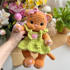 Pode incluir: Uma boneca de gato de croch&ecirc; laranja feita &agrave; m&atilde;o, vestindo um vestido verde claro com um cora&ccedil;&atilde;o rosa e uma faixa de cabelo rosa com um la&ccedil;o. O gato tem almofadas de pata rosa e um nariz rosa. A boneca &eacute; um brinquedo macio.