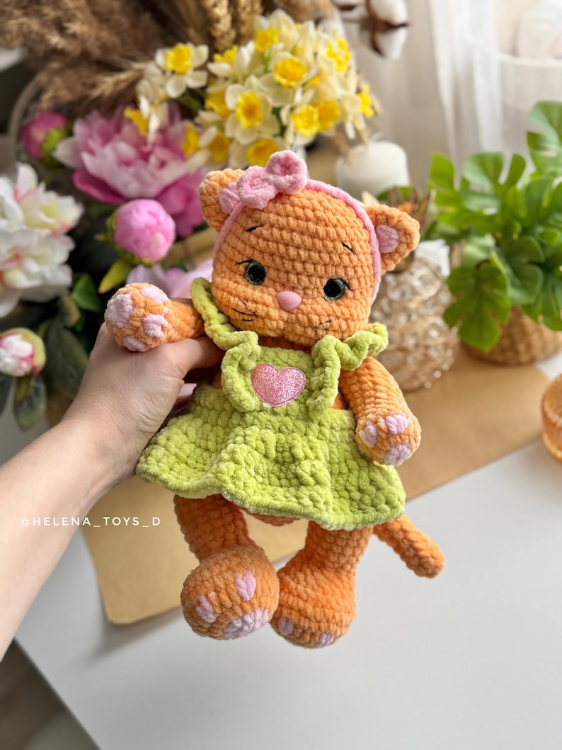 Pode incluir: Uma boneca de gato de croch&ecirc; laranja feita &agrave; m&atilde;o, vestindo um vestido verde claro com um cora&ccedil;&atilde;o rosa e uma faixa de cabelo rosa com um la&ccedil;o. O gato tem almofadas de pata rosa e um nariz rosa. A boneca &eacute; um brinquedo macio.