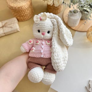 May include: A handmade crochet bunny doll with a white body, long floppy ears, and a pink nose. The bunny wears a pink sweater with flower accents and brown pants. A small bow adorns the top of its head. The doll is soft and cuddly.