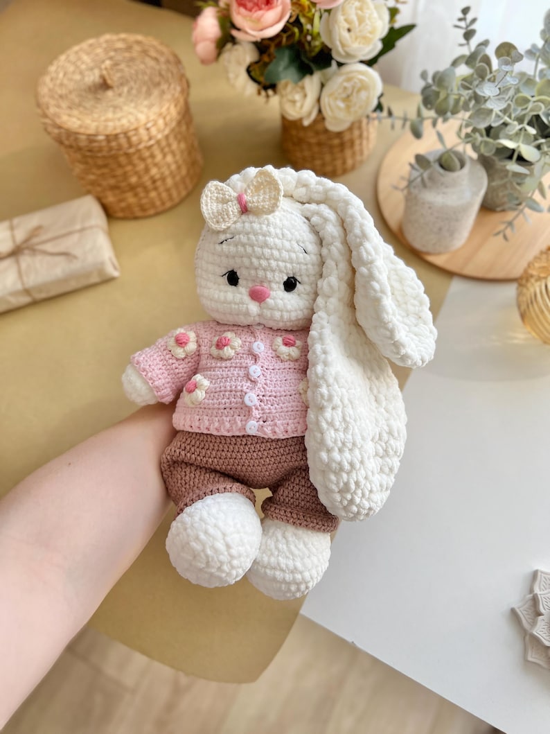 May include: A handmade crochet bunny doll with a white body, long floppy ears, and a pink nose. The bunny wears a pink sweater with flower accents and brown pants. A small bow adorns the top of its head. The doll is soft and cuddly.