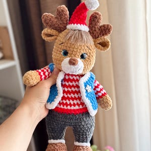 May include: A handmade crochet reindeer doll wearing a red and white striped sweater, blue vest, and gray pants. The reindeer has brown antlers, a red Santa hat with a white pom-pom, and blue eyes. The doll is a festive holiday decoration.
