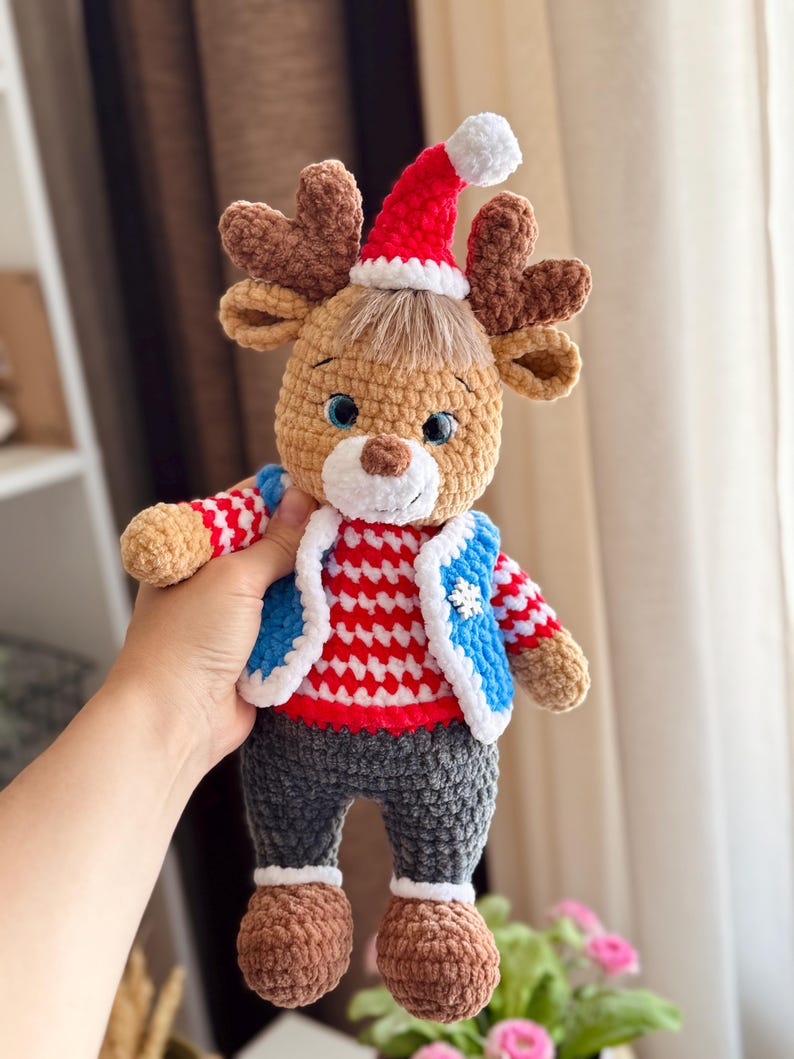 May include: A handmade crochet reindeer doll wearing a red and white striped sweater, blue vest, and gray pants. The reindeer has brown antlers, a red Santa hat with a white pom-pom, and blue eyes. The doll is a festive holiday decoration.