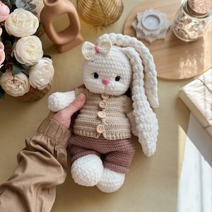 May include: A handmade crochet bunny doll with long, floppy ears. The bunny wears a tan sweater with wooden buttons and brown shorts. It has a pink bow and a pink nose. The doll is white, tan, and brown.