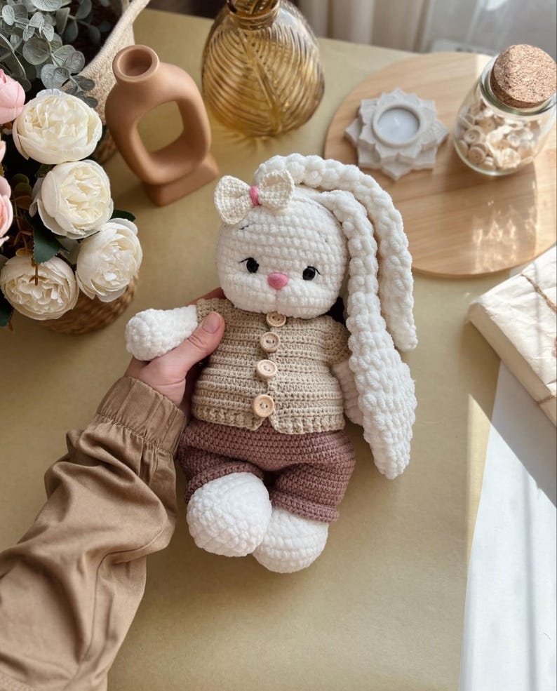May include: A handmade crochet bunny doll with long, floppy ears. The bunny wears a tan sweater with wooden buttons and brown shorts. It has a pink bow and a pink nose. The doll is white, tan, and brown.