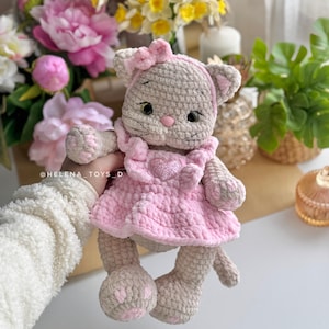 Pode incluir: Boneca de gato de croch&ecirc; feita &agrave; m&atilde;o, vestindo um vestido rosa com um enfeite em forma de cora&ccedil;&atilde;o. A boneca tem um la&ccedil;o rosa e &eacute; cinza claro e bege. O fundo inclui flores e vegeta&ccedil;&atilde;o.