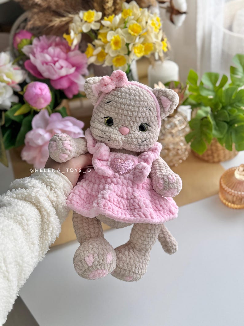 Pode incluir: Boneca de gato de croch&ecirc; feita &agrave; m&atilde;o, vestindo um vestido rosa com um enfeite em forma de cora&ccedil;&atilde;o. A boneca tem um la&ccedil;o rosa e &eacute; cinza claro e bege. O fundo inclui flores e vegeta&ccedil;&atilde;o.