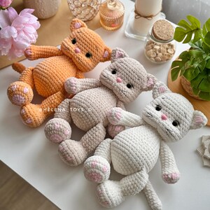 Pode incluir: Tr&ecirc;s pel&uacute;cias de gato de croch&ecirc; em laranja, bege e branco, cada uma com almofadas de pata rosa e detalhes faciais. Os brinquedos est&atilde;o sobre uma superf&iacute;cie branca. O texto "HELENA_TOYS_D" &eacute; vis&iacute;vel.