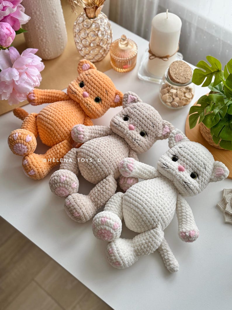 Pode incluir: Tr&ecirc;s pel&uacute;cias de gato de croch&ecirc; em laranja, bege e branco, cada uma com almofadas de pata rosa e detalhes faciais. Os brinquedos est&atilde;o sobre uma superf&iacute;cie branca. O texto "HELENA_TOYS_D" &eacute; vis&iacute;vel.