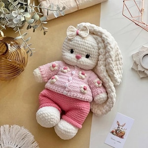 May include: A handmade crochet bunny doll wearing a pink sweater with flower accents and matching pants. The bunny has long, floppy ears, a bow, and white feet. A small card with a bunny illustration is visible.
