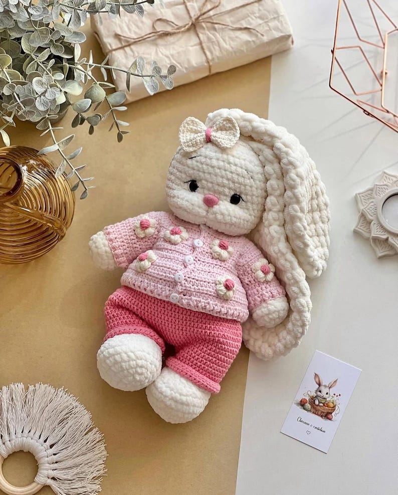 May include: A handmade crochet bunny doll wearing a pink sweater with flower accents and matching pants. The bunny has long, floppy ears, a bow, and white feet. A small card with a bunny illustration is visible.