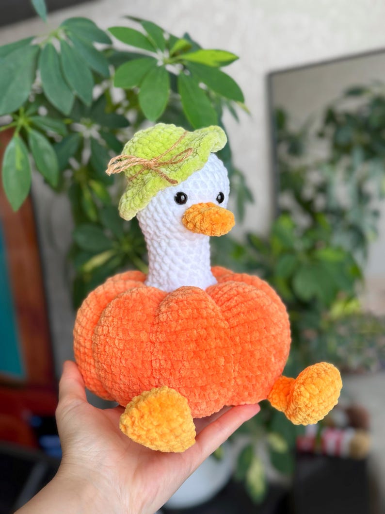 May include: A handmade plush goose with a white head, orange beak, and a green hat. The goose is nestled in an orange pumpkin body with orange feet. The plush toy is made of soft, textured yarn.
