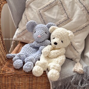 Pode incluir: Dois animais de pel&uacute;cia de croch&ecirc; feitos &agrave; m&atilde;o: um rato azul claro com um nariz rosa e um urso branco. Eles est&atilde;o sentados em uma cadeira de vime com um cobertor cinza e uma almofada decorativa no fundo. Os brinquedos s&atilde;o macios e fofos.