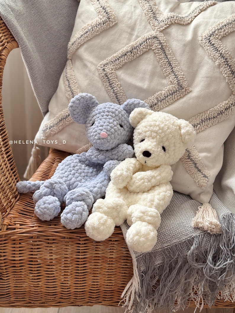 Pode incluir: Dois animais de pel&uacute;cia de croch&ecirc; feitos &agrave; m&atilde;o: um rato azul claro com um nariz rosa e um urso branco. Eles est&atilde;o sentados em uma cadeira de vime com um cobertor cinza e uma almofada decorativa no fundo. Os brinquedos s&atilde;o macios e fofos.