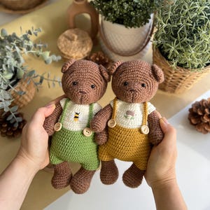 May include: Two handmade crochet teddy bears. One bear wears green overalls with a bee design, the other wears yellow overalls with a flower design. Both bears have brown fur, button accents, and are held by hands.
