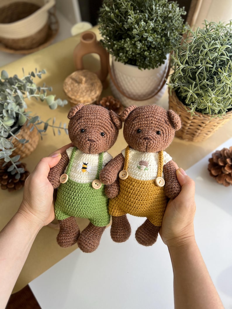 May include: Two handmade crochet teddy bears. One bear wears green overalls with a bee design, the other wears yellow overalls with a flower design. Both bears have brown fur, button accents, and are held by hands.
