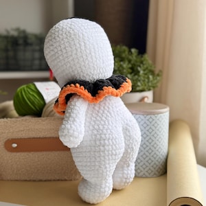 May include: A handmade, white crochet ghost doll with a black and orange ruffled collar. The ghost has a rounded body and is standing on a light brown surface. The doll is a Halloween-themed item.