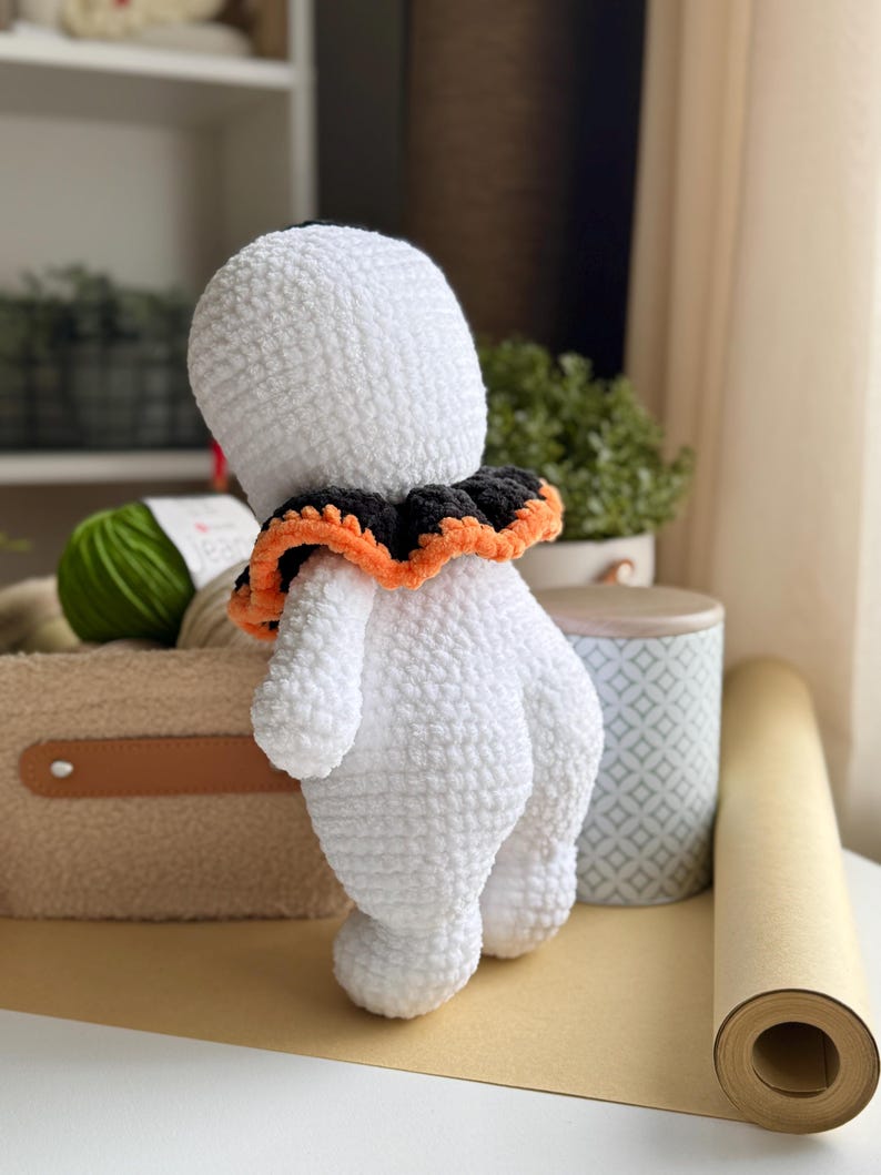 May include: A handmade, white crochet ghost doll with a black and orange ruffled collar. The ghost has a rounded body and is standing on a light brown surface. The doll is a Halloween-themed item.