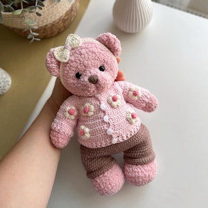 May include: A handmade pink crochet teddy bear wearing a matching sweater with white and pink flower accents and brown pants. The bear has a white bow and button details. The bear is a soft toy.