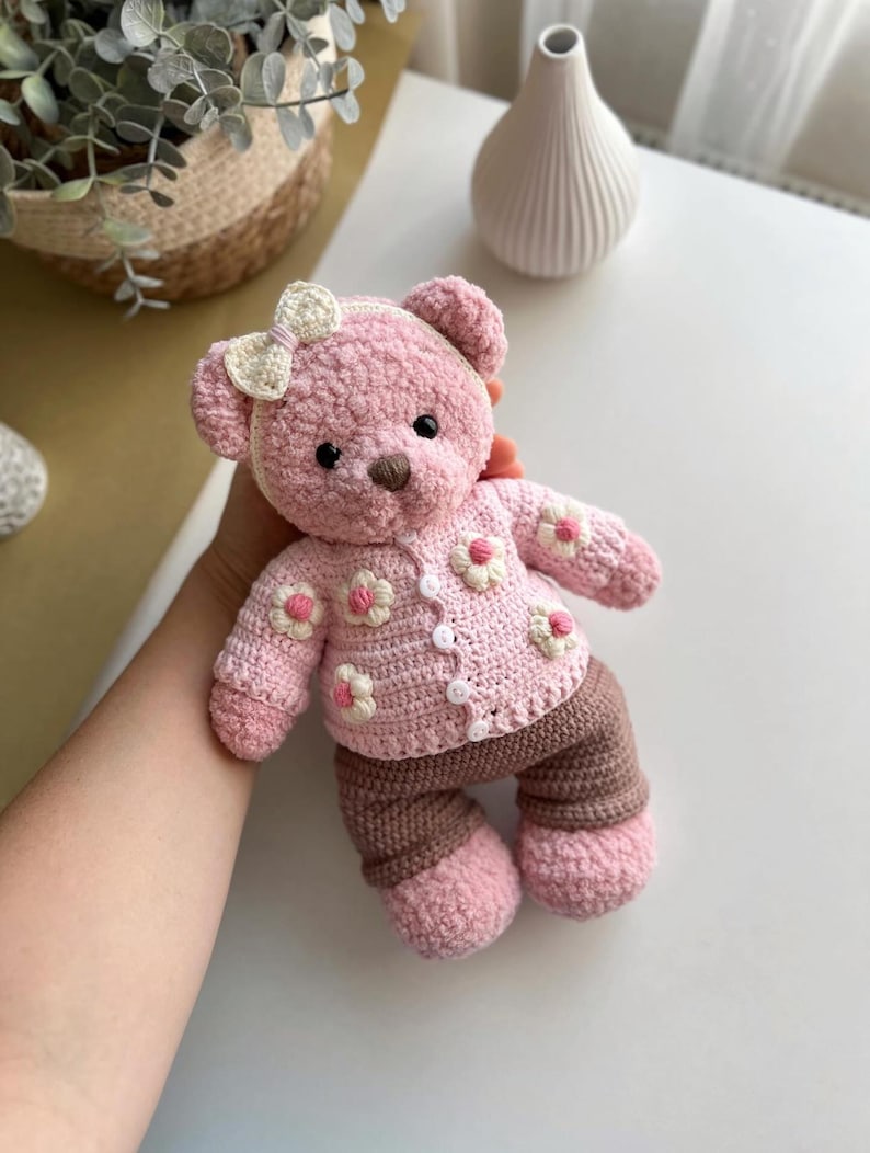 May include: A handmade pink crochet teddy bear wearing a matching sweater with white and pink flower accents and brown pants. The bear has a white bow and button details. The bear is a soft toy.