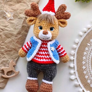 May include: A handmade crochet reindeer doll wearing a red and white striped sweater, blue jacket, and gray pants. The reindeer has brown antlers, a red Santa hat, and brown shoes. A snowflake is on the jacket. Christmas decor.
