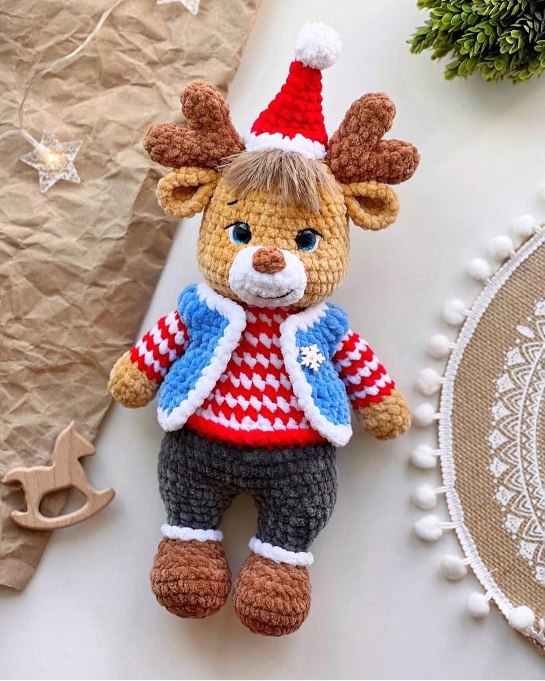 May include: A handmade crochet reindeer doll wearing a red and white striped sweater, blue jacket, and gray pants. The reindeer has brown antlers, a red Santa hat, and brown shoes. A snowflake is on the jacket. Christmas decor.