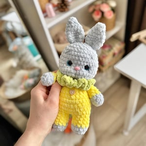May include: A gray crocheted bunny wearing a yellow crocheted outfit with a green ruffle collar. The bunny has a button on its chest.