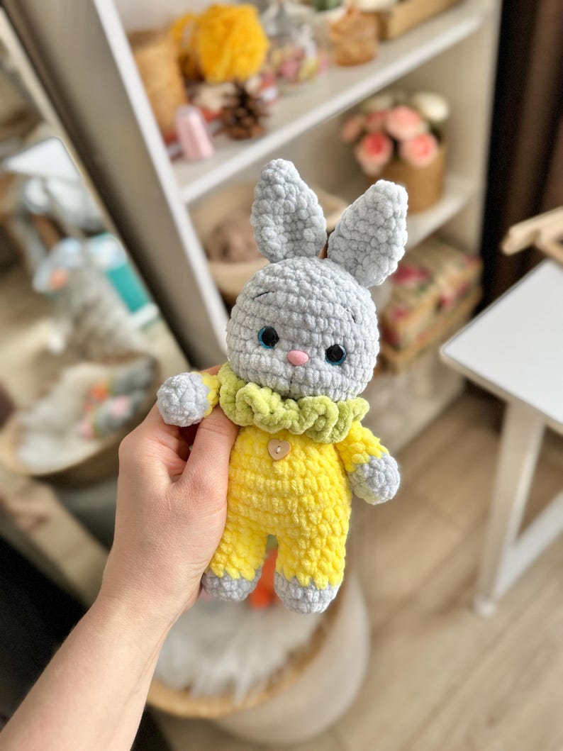 May include: A gray crocheted bunny wearing a yellow crocheted outfit with a green ruffle collar. The bunny has a button on its chest.