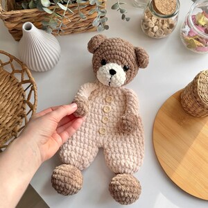 May include: A crocheted teddy bear toy with brown fur and a pink and white outfit. The bear has brown button eyes and a brown nose. The bear is wearing a pink and white outfit with brown buttons. The bear has brown pom-poms on its feet.