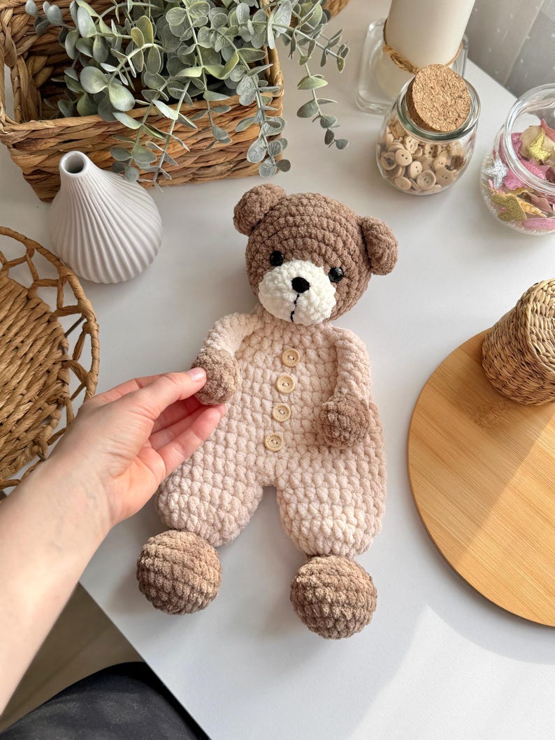 May include: A crocheted teddy bear toy with brown fur and a pink and white outfit. The bear has brown button eyes and a brown nose. The bear is wearing a pink and white outfit with brown buttons. The bear has brown pom-poms on its feet.