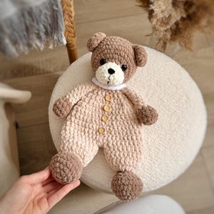 May include: A crocheted brown and beige teddy bear wearing a white collar and brown button accents. The bear is lying on a white fluffy surface.