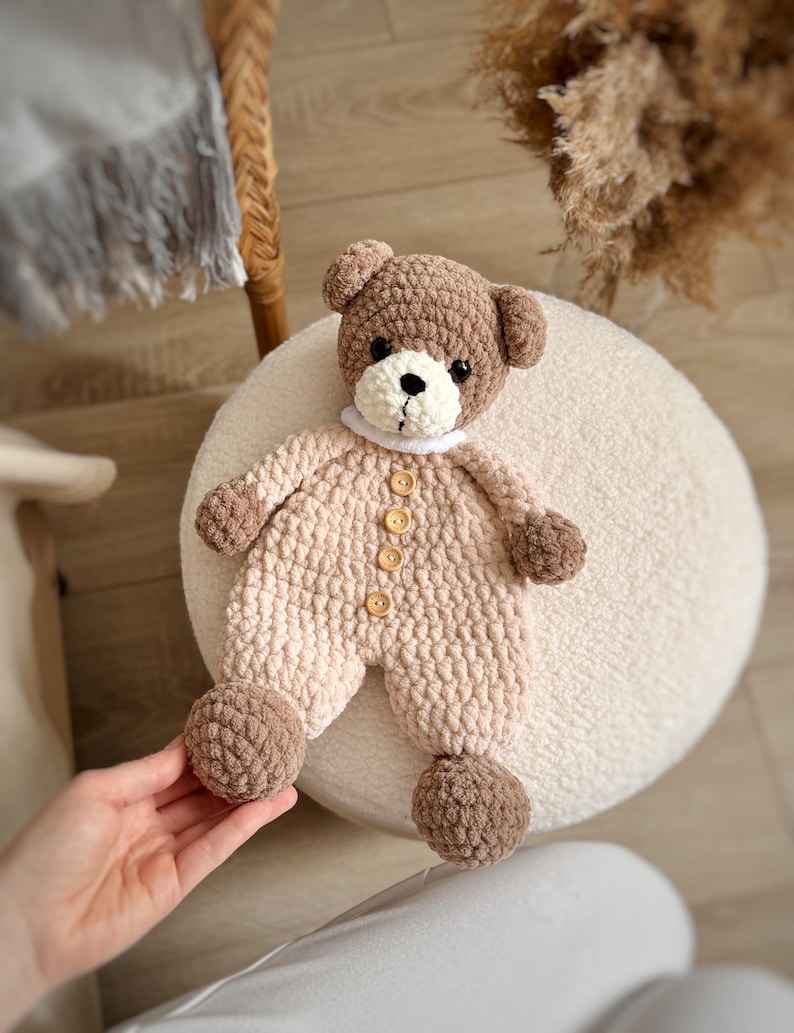May include: A crocheted brown and beige teddy bear wearing a white collar and brown button accents. The bear is lying on a white fluffy surface.
