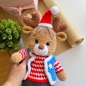 May include: A crocheted reindeer doll wearing a red and white striped sweater, a blue vest with a snowflake, and gray pants. The reindeer has brown antlers, a red Santa hat with a white pom-pom, and blue eyes. A festive holiday decoration.